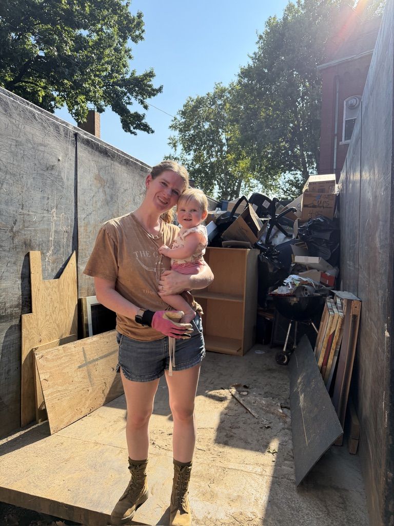 A volunteer in work gloves holding a toddler inside a dumpster full of debris from the church